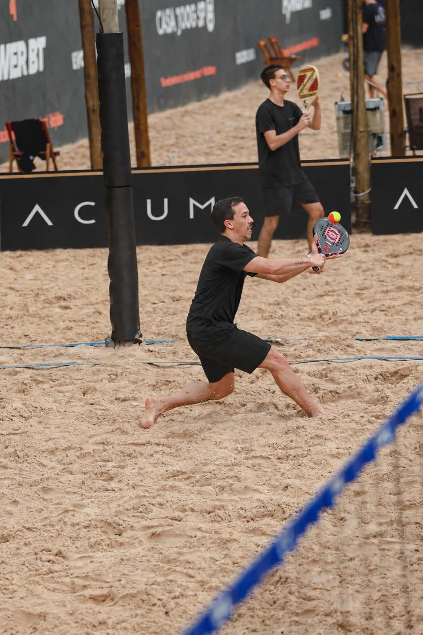 Atleta defende na quadra de areia da Casa JOOG, com placas ACIUM ao fundo